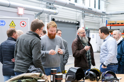 Gruppe von Männern in einer Werkhalle im Gespräch, einige halten Metallteile, auf dem Tisch liegen Schutzausrüstung wie Helme und Schutzbrillen, Warnschilder an der Wand.