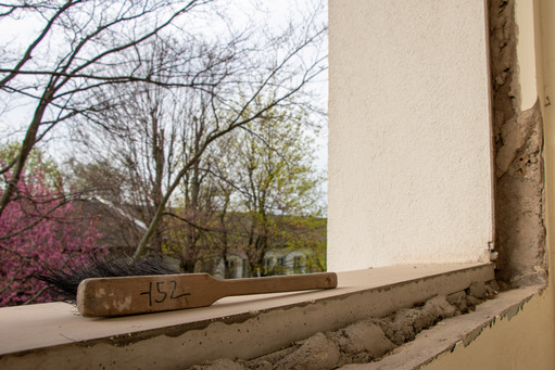 In der Aussparung eines Fensters legt ein alter Handkehrer aus Holz mit der Nummer 752, im Hintergrund lila blühende Bäme