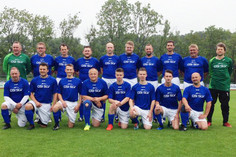 Eine große Gruppe von Männern in Sportkleidung steht und kniet auf einem Fußballfeld, einige tragen blaue Trikots mit SLV Aufdruck, im Vordergrund liegt ein Fußball, im Hintergrund sind Bäume und ein Sportplatz sichtbar