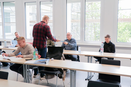 Ein Mann in kariertem Hemd übergibt einem anderen Teilnehmer ein Heft, während andere im Raum sitzen, lesen oder lächeln.