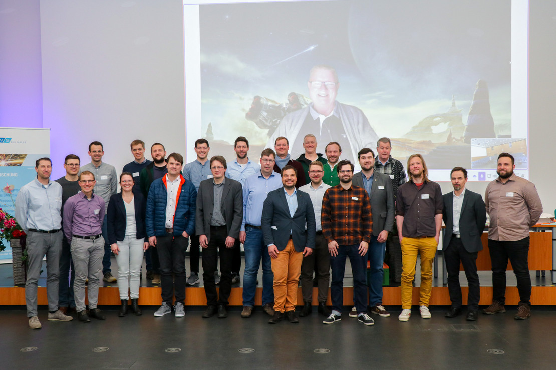 Gruppenfoto mit 27 Personen (25 Männer, 2 Frauen) auf einer Bühne vor einer großen Projektionsfläche mit virtuellem Hintergrund. Eine hybride Konferenzsituation ist erkennbar.