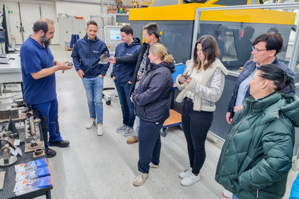 Sieben Personen stehen in einer Werkhalle und hören einem Mann zu, der verschiedene Werkstücke und Schweißteile auf einem Tisch erklärt.