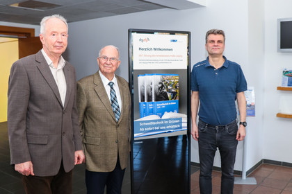 Drei Personen stehen nebeneinander in einem Innenraum vor einer digitalen Stehle, die die SLV-Chronik bewirbt. Links und in der Mitte tragen sie Anzüge, rechts trägt die Person ein kurzes T-Shirt und Jeans.