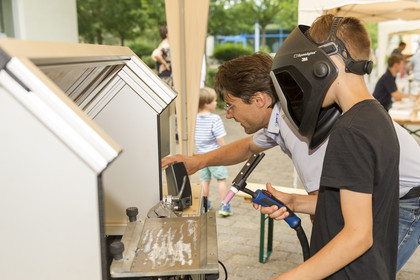 Ein Jugendlicher mit Schweißhelm probiert an einem Schweißsimulator das Schweißen aus, eine begleitende Person unterstützt ihn dabei; im Hintergrund ist ein Kind mit gestreiftem Shirt auf einem Hof zu sehen.