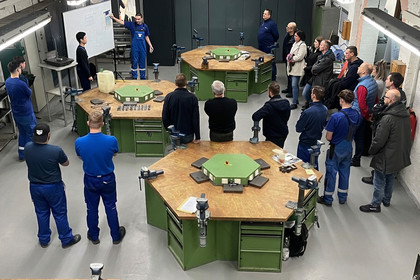 Mehrere Personen stehen in einer Werkhalle um drei große grüne Arbeitstische und hören einer erklärenden Person zu, technische Ausstattung und Maschinen sind im Raum verteilt.