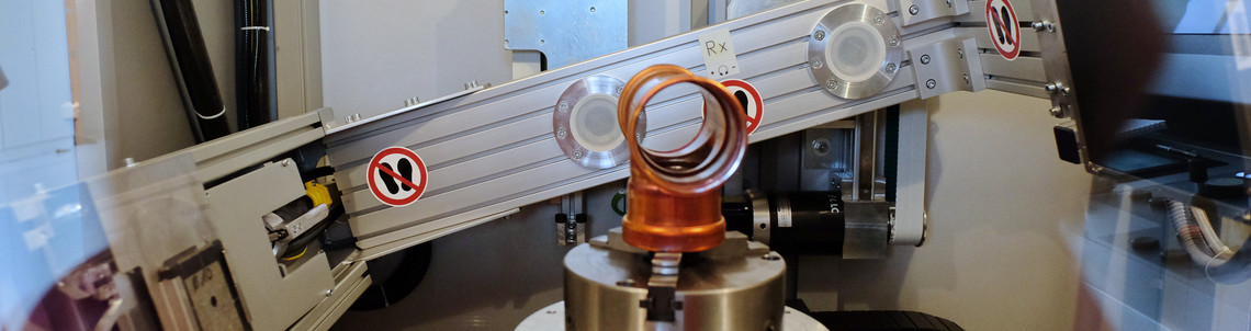 Ein rotationssymmetrisches Bauteil ist in einem Prüfgerät eingespannt, das mit zwei gegenüberliegenden Röntgenquellen oder Detektoreinheiten ausgestattet ist. Die Anlage befindet sich in einem abgeschirmten Raum für zerstörungsfreie Werkstoffprüfung.