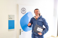 Ein lächelnder Mann mit Brille steht vor einer Tür mit der Aufschrift „Hey, Alter! Alte Rechner für junge Leute“, trägt eine blaue Jacke und hält einen Laptop mit GSI SLV Halle-Aufkleber, mit der anderen Hand macht er ein Daumen-Hoch-Zeichen
