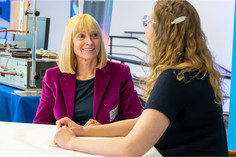 Zwei Frauen sitzen sich an einem Tisch gegenüber und führen ein Gespräch, beide hören aufmerksam zu, im Hintergrund sind Ausstellungsstände der Schweißtechnischen Tagung zu sehen.