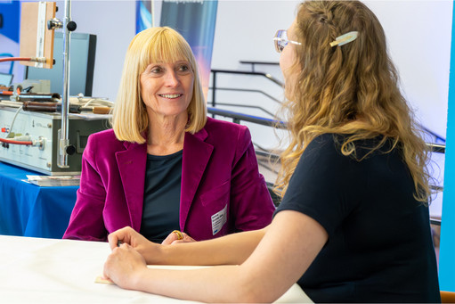 Zwei Frauen sitzen sich an einem Tisch gegenüber und führen ein Gespräch, beide hören aufmerksam zu, im Hintergrund sind Ausstellungsstände der Schweißtechnischen Tagung zu sehen.