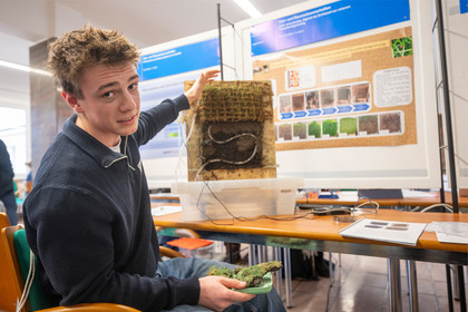 Jugendlicher sitzt an einem Arbeitstisch vor einer geöffneten elektronischen Baugruppe, die mit Rasen verbunden ist, und blickt zur Kamera, im Hintergrund ist ein Informationsplakat mit technischen Grafiken und Gebäudedarstellungen zu sehen.