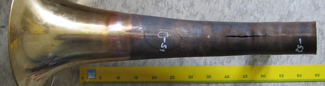 Ein beschädigter Blasinstrument-Schalltrichter liegt auf dem Boden, das Material zeigt einen langen Längsriss und ist mit Messmarkierungen versehen, daneben liegt ein Maßband.