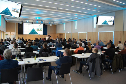 Großer Konferenzraum mit zahlreichen Teilnehmenden an Tischen, alle blicken zur Bühne mit Präsentationsleinwand und Redner, moderne Beleuchtung und mehrere Bildschirme an den Wänden