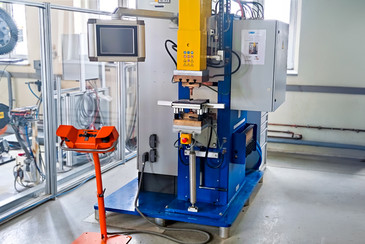 Eine industrielle Punktschweißmaschine mit gelbem Oberteil und blauem Sockel steht in einer Werkhalle; daneben befindet sich ein orangefarbenes Bedienpult auf Ständer.