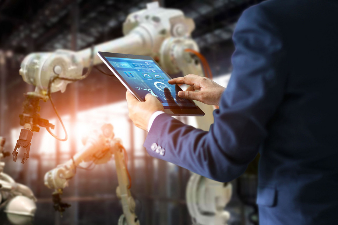 Mann im Anzug bedient ein Tablet mit digitalen Steuerungsanzeigen, im Hintergrund arbeiten mehrere Industrieroboter.