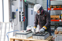 Ein Mann bearbeitet mit einem handgeführten Werkzeug eine Batterie auf einem Arbeitstisch in einer Werkhalle. Er trägt einen weißen Schutzhelm mit Visier und Schutzhandschuhe, im Hintergrund sind Regale und weitere Arbeitsgeräte zu sehen.