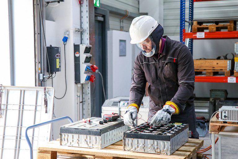 Ein Mann bearbeitet mit einem handgeführten Werkzeug eine Batterie auf einem Arbeitstisch in einer Werkhalle. Er trägt einen weißen Schutzhelm mit Visier und Schutzhandschuhe, im Hintergrund sind Regale und weitere Arbeitsgeräte zu sehen.