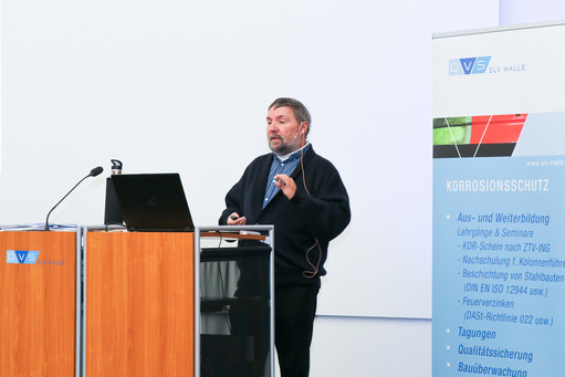 Ein Mann steht an einem Rednerpult und hält einen Vortrag mit Mikrofon vor einem Rollup Banner und einer Präsentationsfläche in einem Veranstaltungsraum.