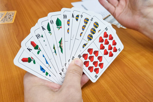 Zwei Hände halten aufgefächerte Spielkarten mit traditionellen Symbolen über einem Holztisch.