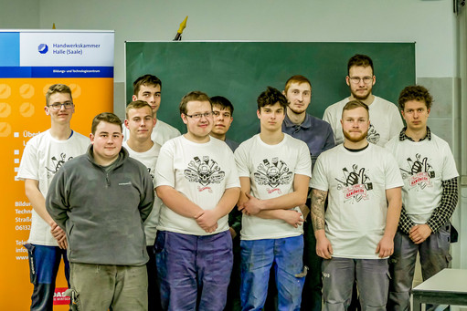 Elf junge Männer stehen in zwei Reihen vor einer Tafel, zehn tragen weiße T-Shirts mit Werkzeugmotiv, einer grauen Pullover; rechts Banner der Handwerkskammer Halle (Saale)