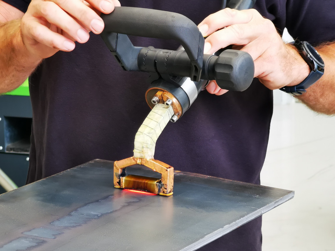 Eine Person hält ein Handgerät zum induktiven Richten über eine Metallplatte mit Schweißnaht; das Gerät leuchtet rot an der Kontaktstelle.