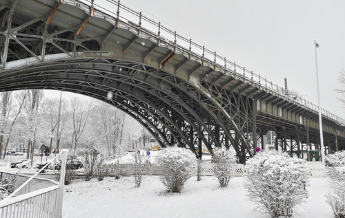 Eine große Brücke aus Altstahl überspannt eine schneebedeckte Landschaft. Der Boden ist vollständig mit Schnee bedeckt, vereinzelt stehen kleine Büsche oder Bäume im Vordergrund.