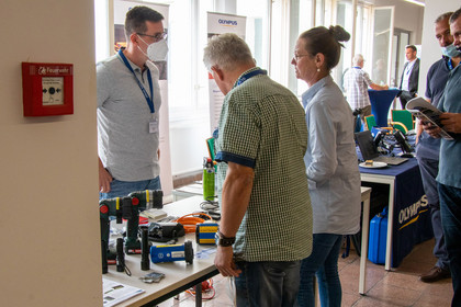 Mehrere Personen stehen an einem Messestand mit Prüfgeräten und technischen Messinstrumenten, ein Mann mit Maske erklärt einem interessierten Paar die ausgestellten Geräte.