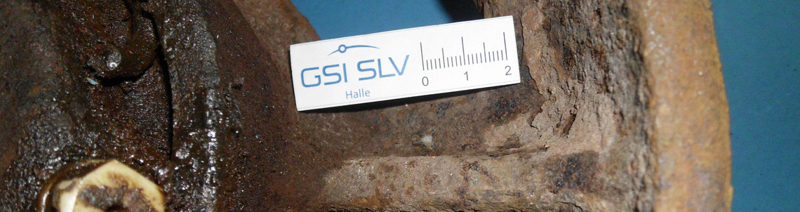 Nahaufnahme eines stark korrodierten Gussrades mit deutlichem Riss entlang einer Speiche, daneben liegt eine Maßskala mit dem Logo der GSI SLV Halle.