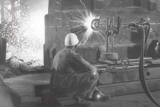 Eine Person mit Schutzhelm sitzt in einer Werkhalle und beobachtet eine Maschine beim thermischen Schneiden eines großen Werkstücks. Funken sprühen hell auf, die Szene ist in Schwarz-Weiß gehalten.