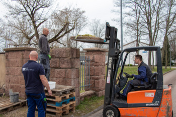 Drei Männer arbeiten im Freien an einer Steinmauer, einer steht auf Paletten, ein anderer bedient einen Gabelstapler mit Material auf der Gabel, alle tragen Arbeitskleidung, im Hintergrund Bäume ohne Laub.