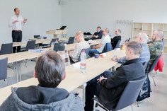 Der Dozent hält eine DVS Urkunde hoch und spricht in einem Schulungsraum vor neun zuhörenden Teilnehmenden, die an Tischen sitzen, darunter eine Person in Bundeswehruniform.