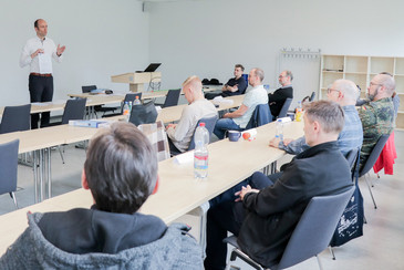 Der Dozent hält eine DVS Urkunde hoch und spricht in einem Schulungsraum vor neun zuhörenden Teilnehmenden, die an Tischen sitzen, darunter eine Person in Bundeswehruniform.