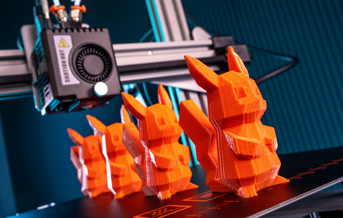 Ein Roboterarm druckt mehrere orangefarbene Plastikfiguren an einem industriellen Arbeitsplatz. Im Hintergrund sind weitere Maschinenteile und Vorrichtungen zu sehen.