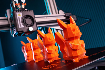 Ein Roboterarm druckt mehrere orangefarbene Plastikfiguren an einem industriellen Arbeitsplatz. Im Hintergrund sind weitere Maschinenteile und Vorrichtungen zu sehen.