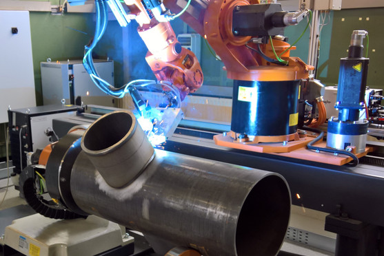 Ein Schweißroboter bearbeitet automatisiert eine Rohr-T-Verbindung in einer Fertigungszelle; das Werkstück ist eingespannt und die Lichtbogenentladung gut sichtbar.