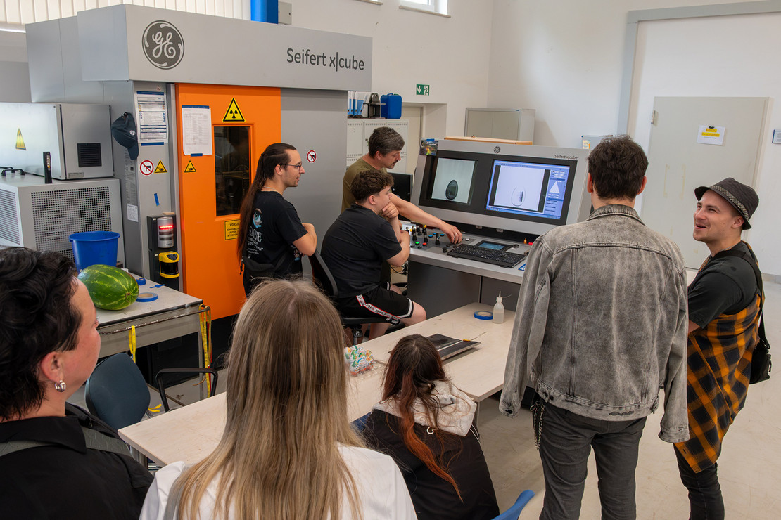 Sieben Personen, darunter Frauen und Männer, stehen in einer Werkhalle vor mehreren Bildschirmen und einer industriellen Anlage. Eine Person zeigt auf die Monitore, während die Gruppe die dargestellten Informationen und die technische Ausrüstung betrachtet.