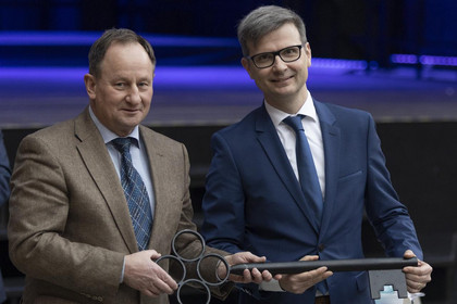 Zwei Männer im Anzug stehen nebeneinander und halten gemeinsam einen übergroßen symbolischen Schlüssel; im Hintergrund eine Bühne mit blauem Licht.