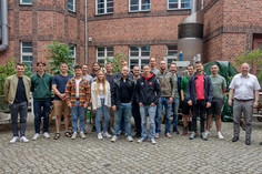 Gruppenfoto auf einem gepflasterten Innenhof vor einem Backsteingebäude mit technischen Anlagen und Pflanzen.