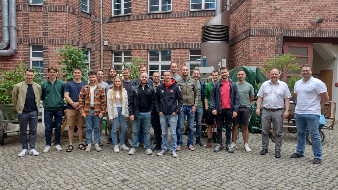 Gruppenfoto auf einem gepflasterten Innenhof vor einem Backsteingebäude mit technischen Anlagen und Pflanzen.