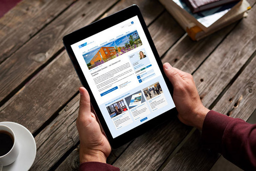 Ein Mann hält ein Tablet mit der Webseite der Betriebsstätte Dresden in der Hand, daneben steht eine Tasse Kaffee. Das Bild ist aus der Vogelperspektive aufgenommen.