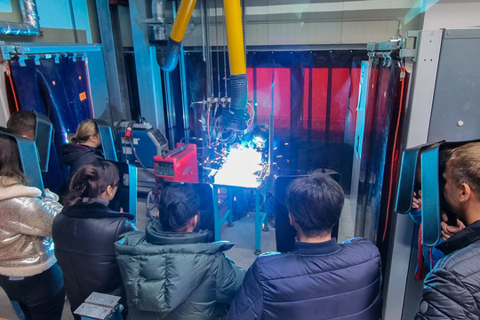 Mehrere Personen beobachten in einer Werkstatt eine Schweißvorführung, bei der ein heller Lichtbogen sichtbar ist, umgeben von Schweißschutzvorhängen.