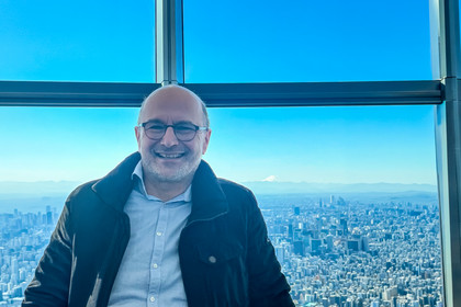 Ein Mann mit Brille steht vor einer großen Fensterfront und blickt in die Kamera. Im Hintergrund ist eine dicht bebaute Stadtlandschaft aus großer Höhe zu sehen, am Horizont zeichnet sich ein Berg ab.