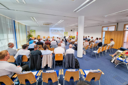 Mehrere Personen sitzen in einem Seminarraum auf Stühlen und blicken auf eine Projektionsfläche mit der Aufschrift „Herzlich Willkommen!“. Vorne stehen ein Rednerpult, technische Geräte und Roll-up-Banner mit Bezug zum Schienenverkehr.