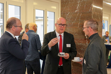 Drei Männer im Anzug unterhalten sich bei einer Veranstaltung in einem hellen Raum mit Marmorsäulen und großen Fenstern; zwei von ihnen halten Kaffeetassen.