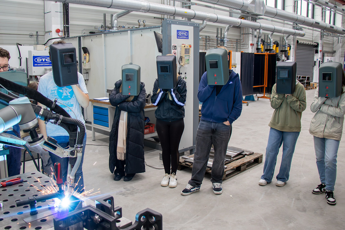 Eine Gruppe von Schülern steht mit Schweißhelmen vor einem Roboter, der aktiv Schweißarbeiten durchführt.