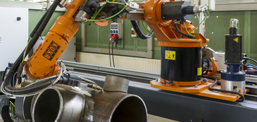 Ein orangefarbener Industrieroboter bearbeitet in einer Werkhalle ein komplex verschweißtes Rohrbauteil auf einer Lineareinheit, umgeben von Kabeln und Steuerungselementen.