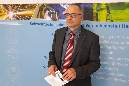 Ein Mann im Anzug mit rot-gestreifter Krawatte steht vor einer blauen Infowand der SLV Halle; er hält ein gefaltetes Informationsblatt in der Hand und blickt zur Seite.