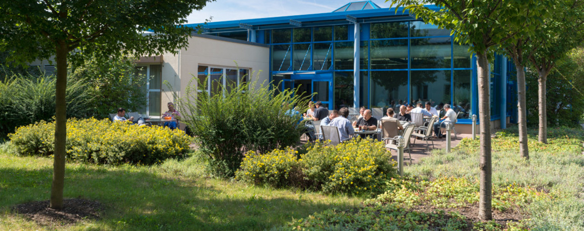 Mehrere Personen sitzen bei sonnigem Wetter auf einer begrünten Terrasse vor einem modernen Gebäude mit blauen Stahl-Glas-Elementen. Die Szene vermittelt eine entspannte Atmosphäre während einer Pause oder Veranstaltung im Grünen.