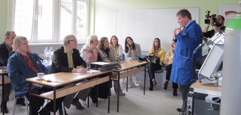 Ein Mann im blauen Kittel spricht vor einer Gruppe von Erwachsenen, darunter mehrere Frauen und Männer, in einem Schulungsraum mit grünen Wänden und Fenstern, im Vordergrund steht ein Schweißsimulator.