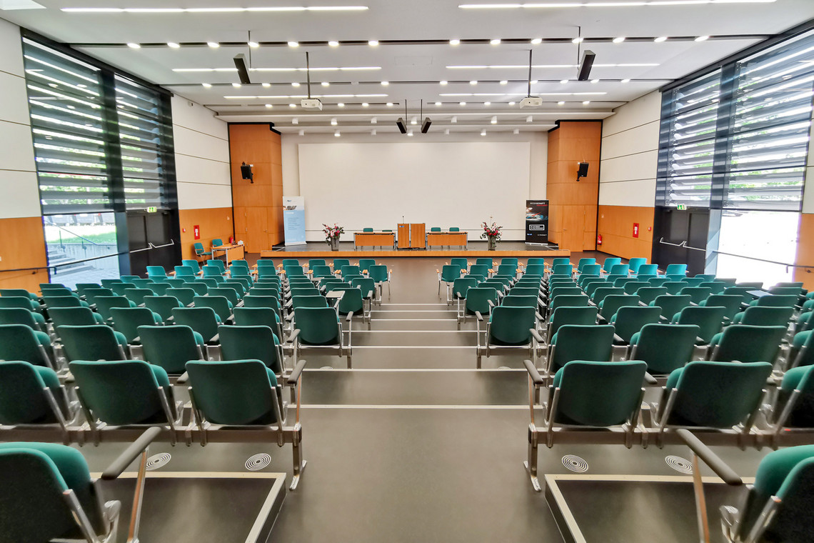 Ein großer Konferenzsaal mit grünen Stuhlreihen, einer großen Leinwand und einem Rednerpult im Vordergrund. Der Raum ist lichtdurchflutet.