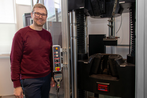 Ein Mann mit Brille und bordeauxfarbenem Pullover steht lächelnd neben einer Prüfmaschine der Marke MTS mit Bedienpanel; im Hintergrund ein helles Labor mit Tageslicht.